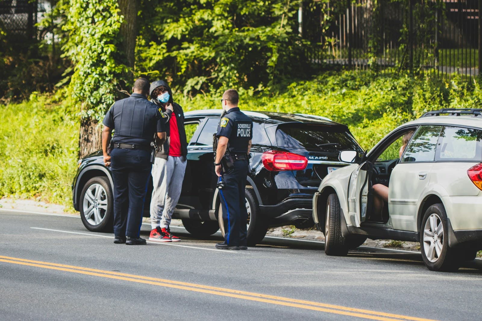 Police officers conducting a traffic stop with a person next to a dark SUV on a tree-lined road